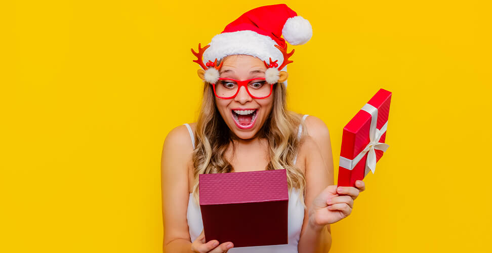 Woman in Santa hat and Christmas glasses opening a festive gift with a shocked happy face &ndash; holiday celebration, Christmas surprise present moment