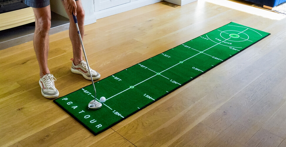 PGA Tour 8FT Deluxe Putting Mat on wooden floor with golfer’s legs and putter ready to hit the ball
