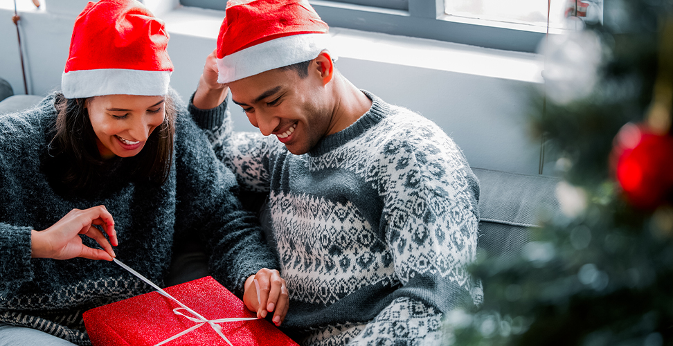 Couple in Santa hats cuddling on sofa by Christmas tree as woman opens festive holiday gift