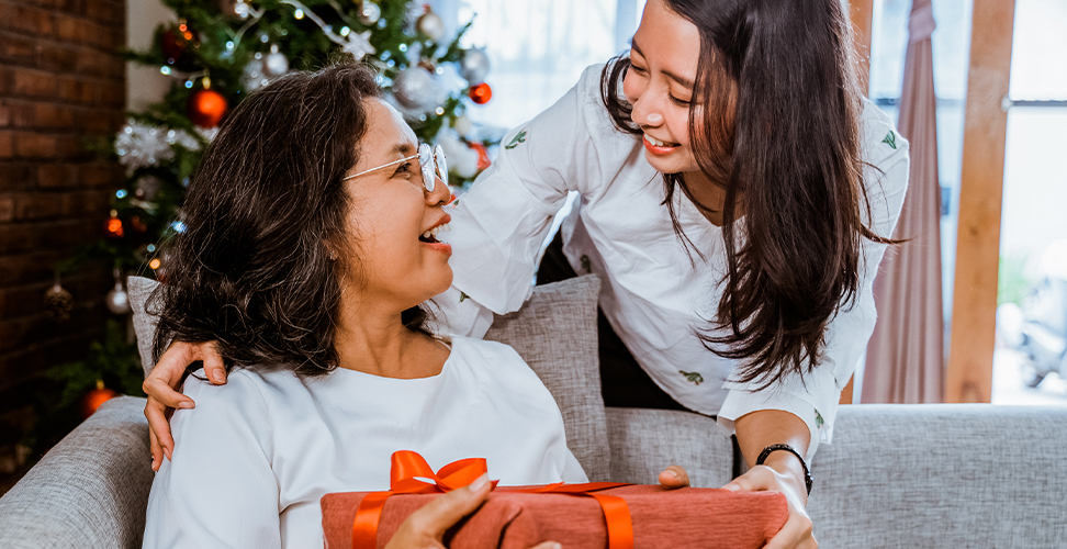 Daughter giving Christmas gift to her smiling mum on sofa with festive Christmas tree in background