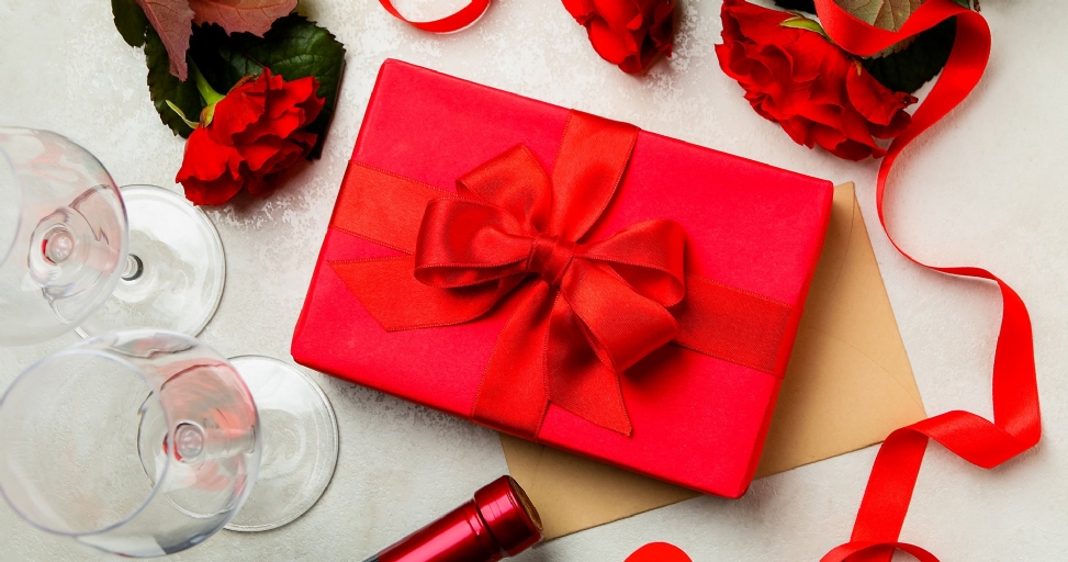 A red gift box with a bow, red roses, wine glasses, and a bottle on a light surface.