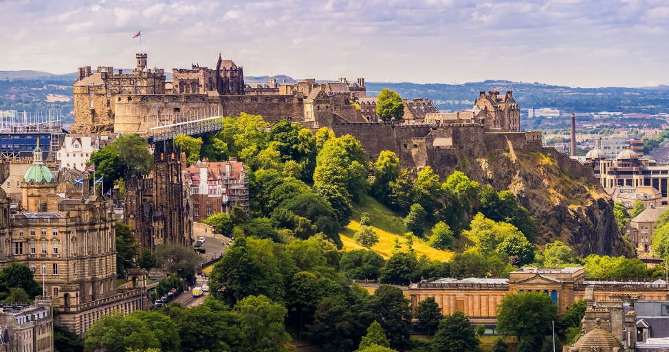 Panoramic view of Edinburgh, Scotland, featuring the historic Edinburgh Castle on a rocky hill amidst lush trees and city buildings.