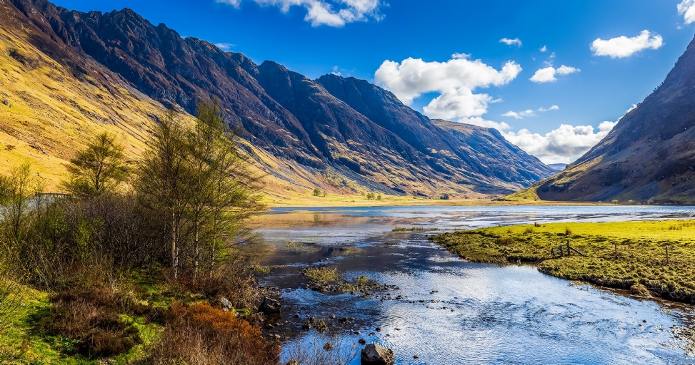 A majestic Scottish glen with towering mountains, a tranquil lake, and a flowing river under a bright sky.