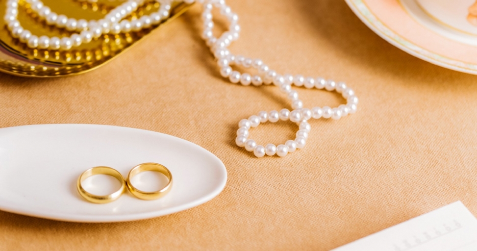 Two gold wedding bands on a white dish, with pearl necklaces on a gold tray and a tan cloth.