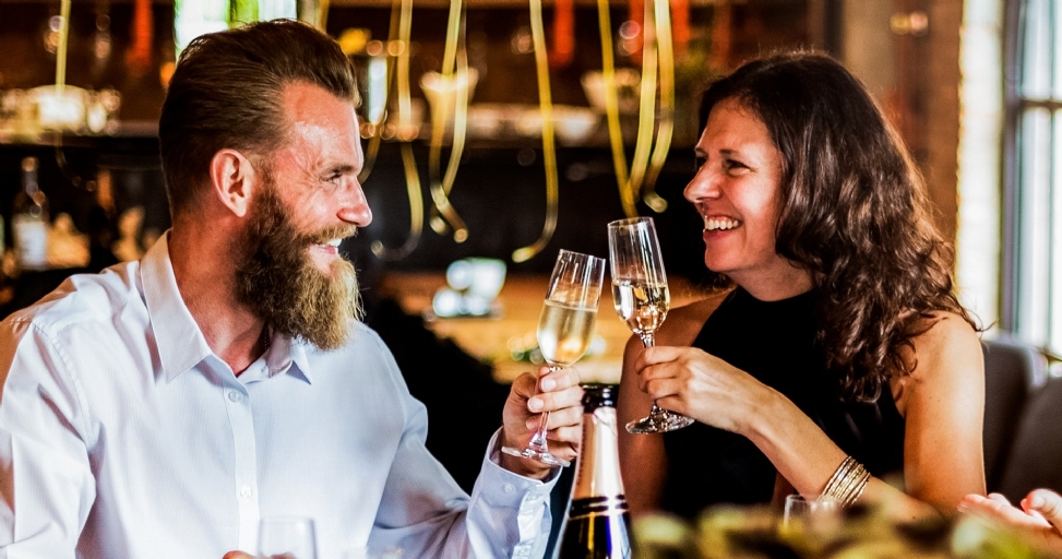 A happy couple smiles and toasts with champagne in a dimly lit restaurant or bar setting.