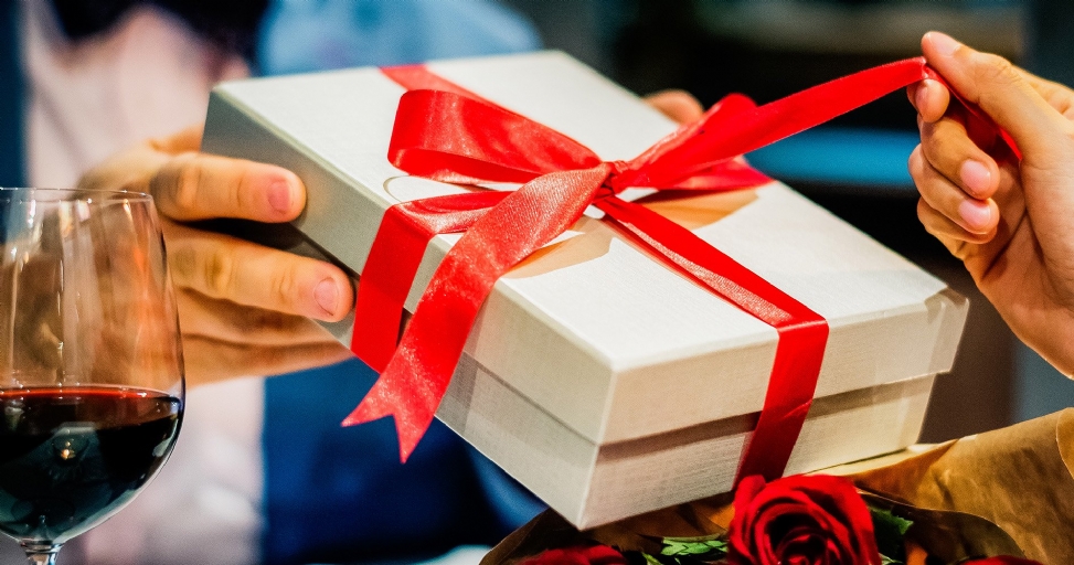 Hands exchanging a white gift box with a red ribbon, with wine and red roses nearby.