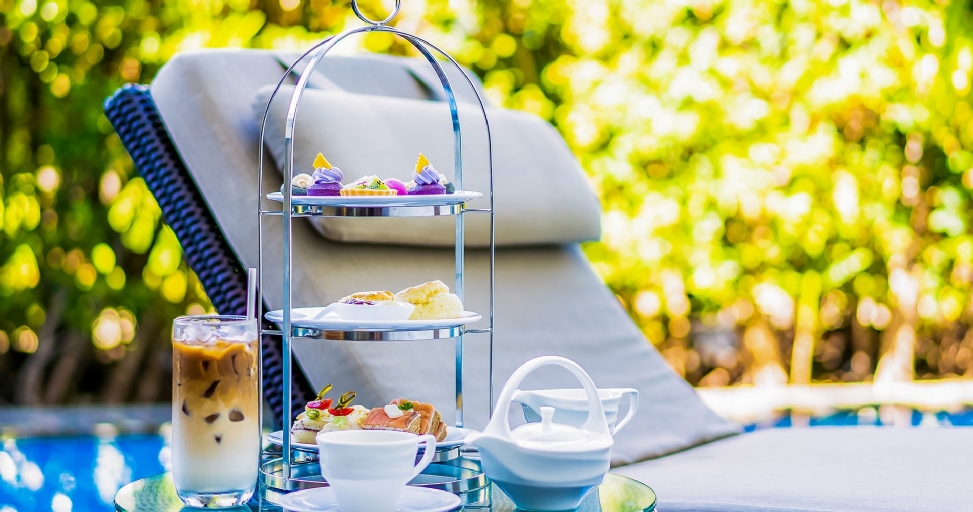 An elegant afternoon tea setup by a pool with a tiered stand of sweets and savories, iced coffee, and a teapot.