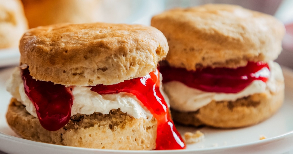 Two delicious golden scones filled with whipped cream and vibrant red jam, with one dripping down.
