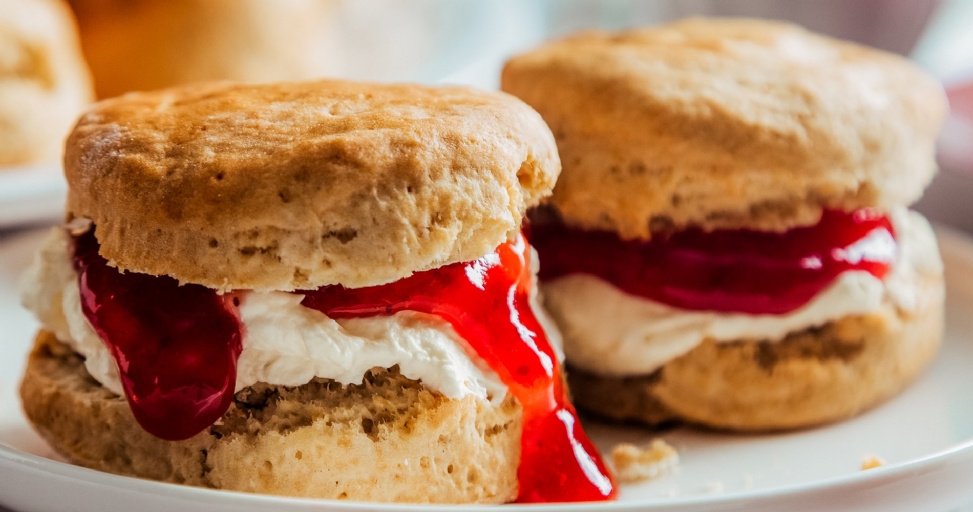 Two delicious scones with white clotted cream and vibrant red jam overflowing on a white plate.