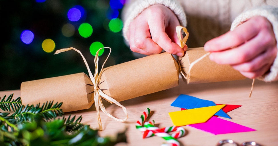 Hands tie raffia around a brown Christmas cracker, surrounded by festive decorations on a wooden table.
