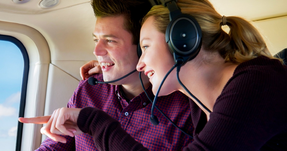 A happy couple in a helicopter, wearing headsets, looking out the window, with the woman pointing.