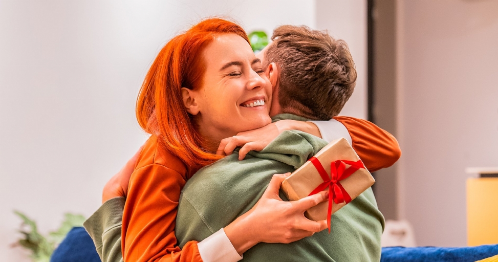 A happy red-haired woman embraces a man holding a gift, both smiling warmly.