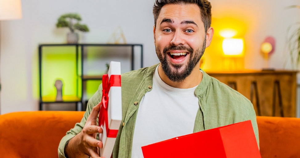 A delighted man with a beard opens a gift box, smiling excitedly at the camera.