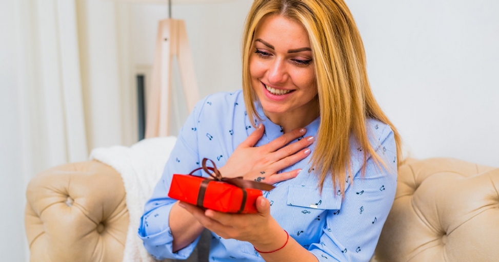 Smiling blonde woman in a blue shirt, looking delighted as she holds a small red gift box.