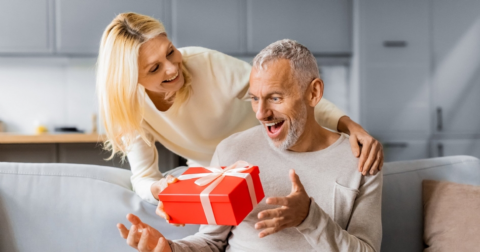 A happy blonde woman gives a red gift box to an excited man on a couch.