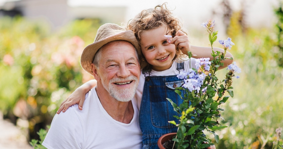 Happy grandfather and granddaughter smiling in a sunny garden, holding a potted blue flowering plant.
