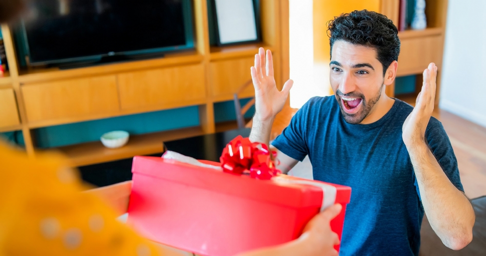 A man reacts with joy and surprise, hands raised, while receiving a red gift box.