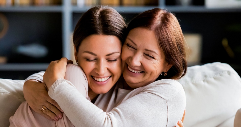 Happy mother and adult daughter hugging, laughing with eyes closed on a couch.