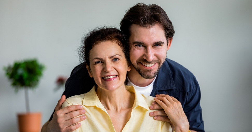 Smiling adult son embracing his joyful mother from behind, both looking at the camera.