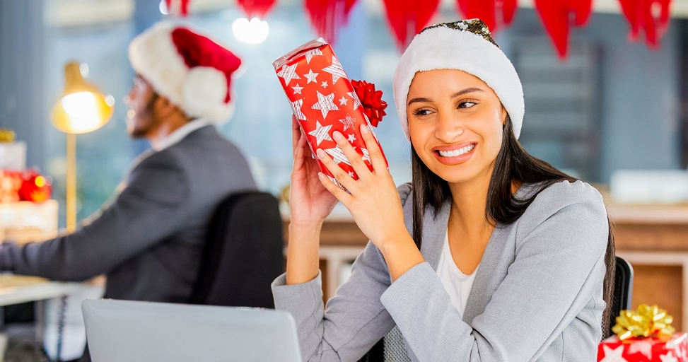 A smiling woman in a Santa hat holds a Christmas gift in a festive office.