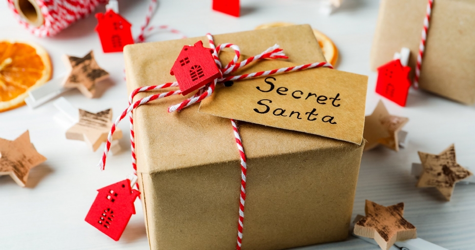 A brown paper gift box with red and white twine, a "Secret Santa" tag, and a red house ornament, surrounded by festive decorations.