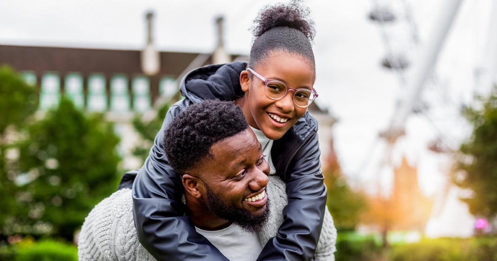A happy Black father gives his smiling daughter a piggyback ride in a park setting.