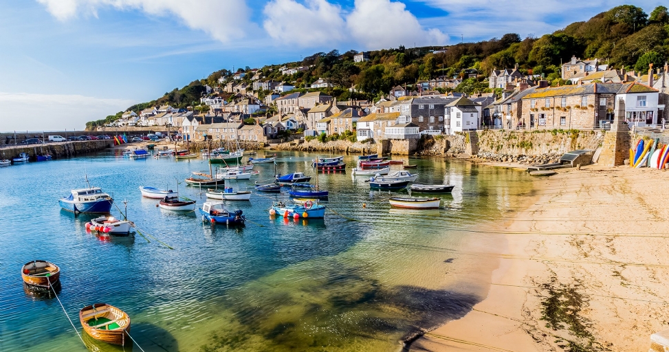 A picturesque coastal village harbor filled with boats, a sandy beach, and hillside houses under a blue sky.