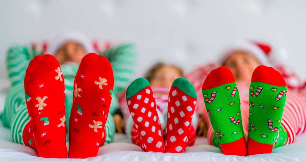 A family's feet, including two adults and a child, wearing festive Christmas socks and pajamas, on a white bed.