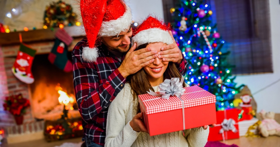 A happy couple in Santa hats celebrating Christmas, with the man surprising the woman with a gift.