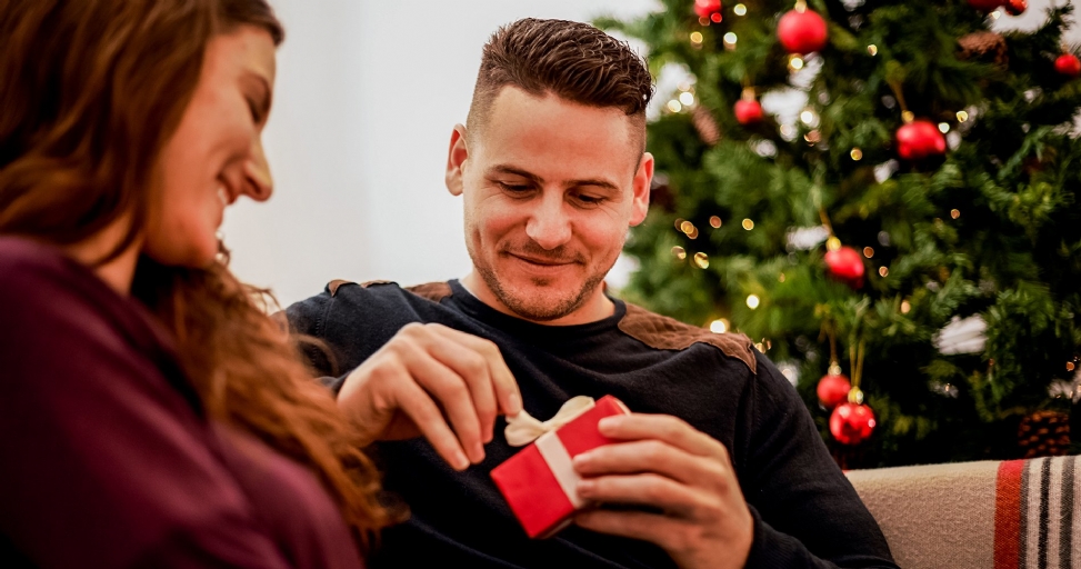 A smiling man unties a white ribbon on a small red gift box, with a woman and Christmas tree nearby.