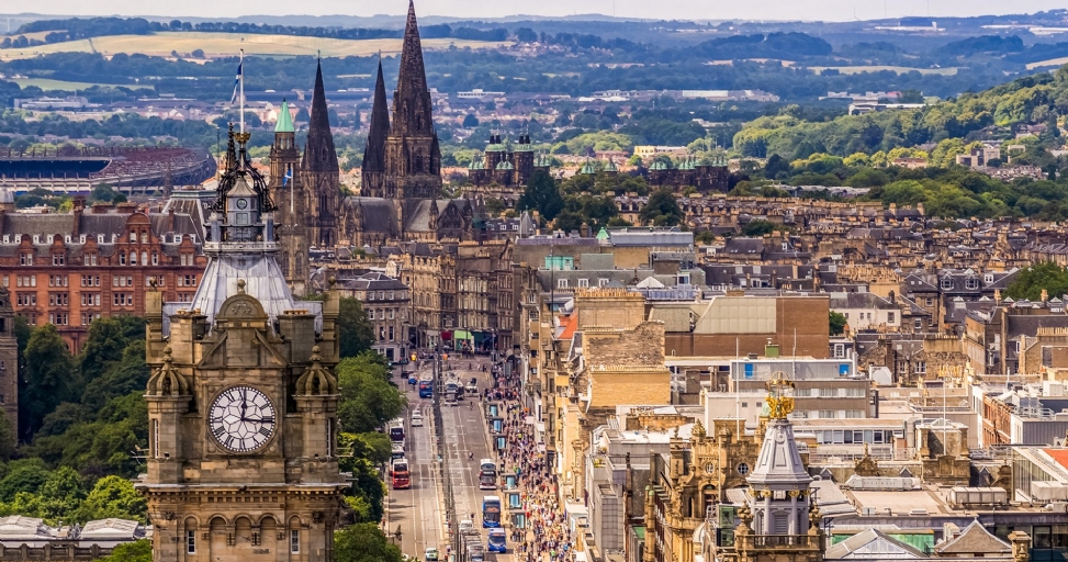 An elevated view of Edinburgh, Scotland, featuring a prominent clock tower, historic buildings, a busy street, and distant green hills.