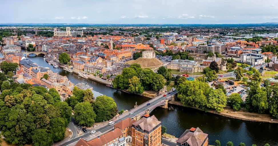 Aerial view of historic York city with a river, bridges, buildings, and green spaces under a cloudy sky.