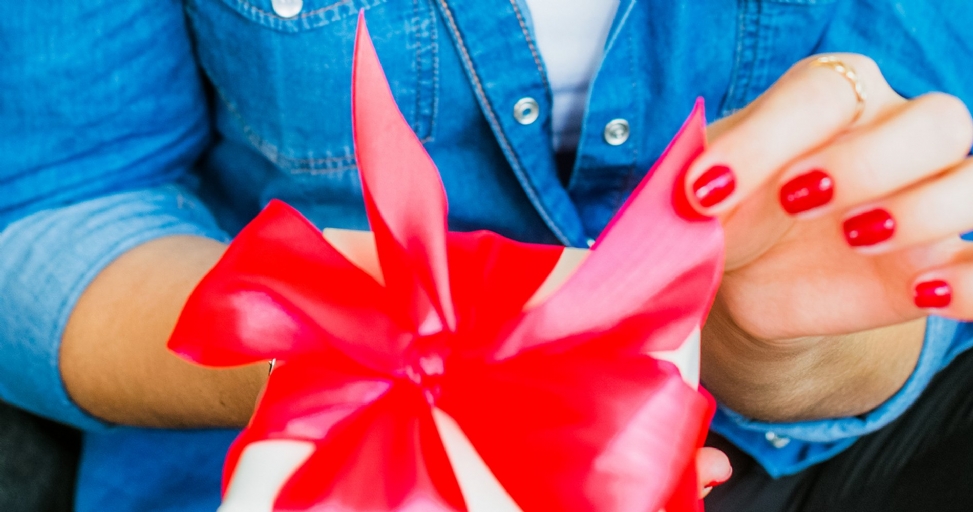 A person's hands with bright red nail polish are opening a gift box with a large red ribbon.