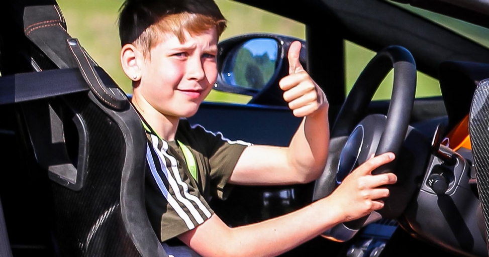 A young boy in a car's driver seat gives a thumbs-up, looking happy and excited.