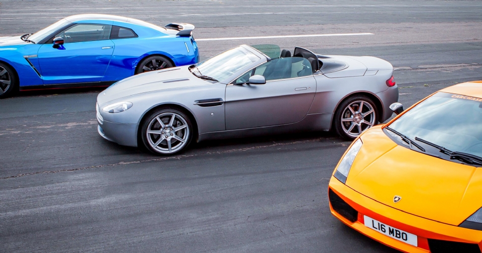 Three luxury sports cars: a blue Nissan GT-R, silver Aston Martin, and orange Lamborghini, parked on asphalt.