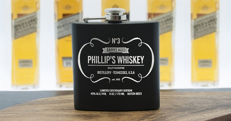 A black engraved flask for whiskey with "Phillip's Whiskey" on a wooden surface with blurred bottles in the background.