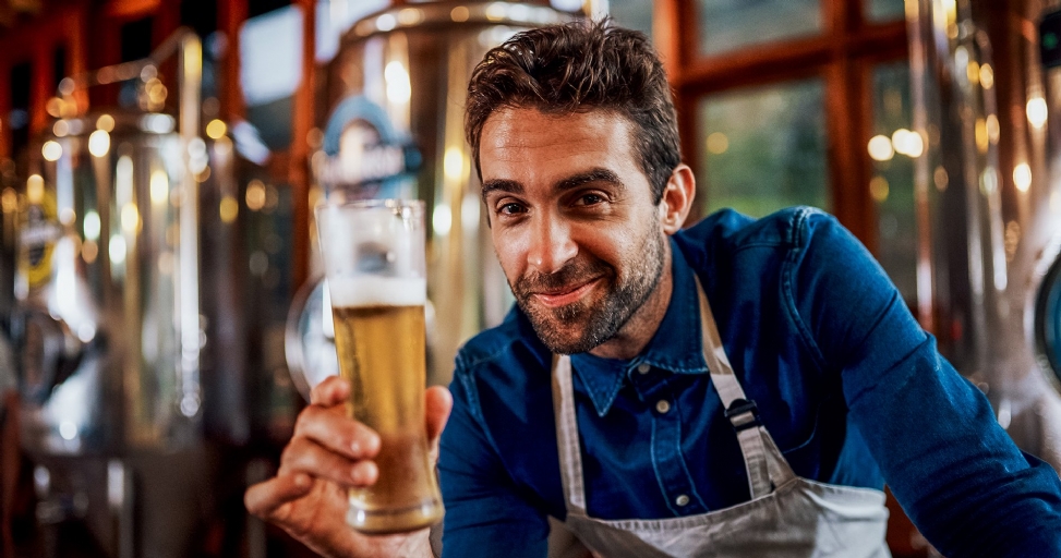 A smiling man in a blue shirt and apron holds out a glass of beer in a brewery.
