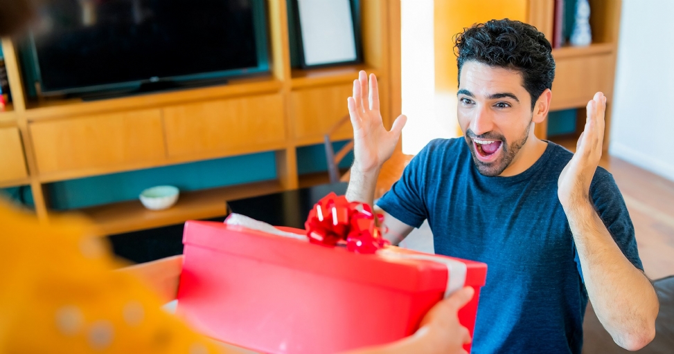 A man looks surprised and happy, hands raised, receiving a red gift box.
