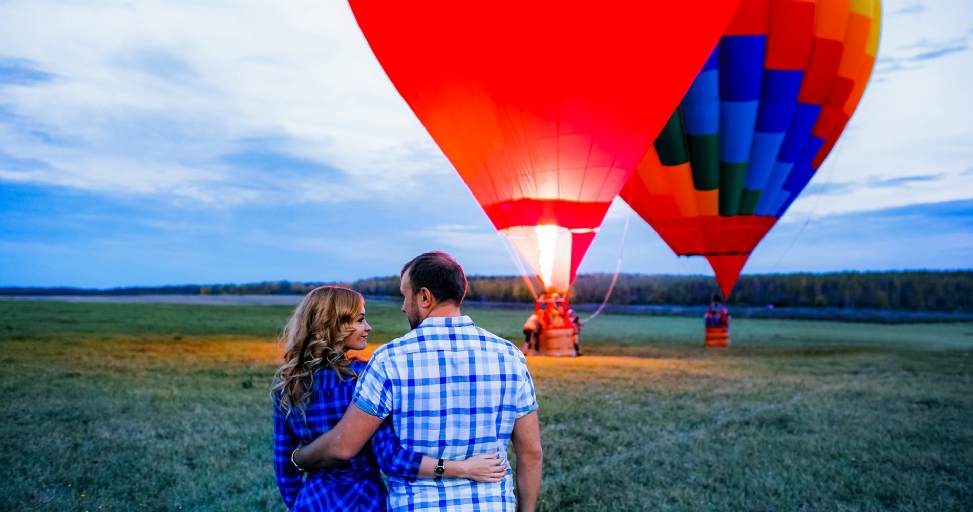 A couple embraces in a field, watching two hot air balloons inflate under a cloudy sky.