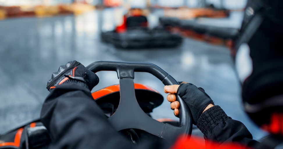 Person's gloved hands on a go-kart steering wheel at an indoor racing track.