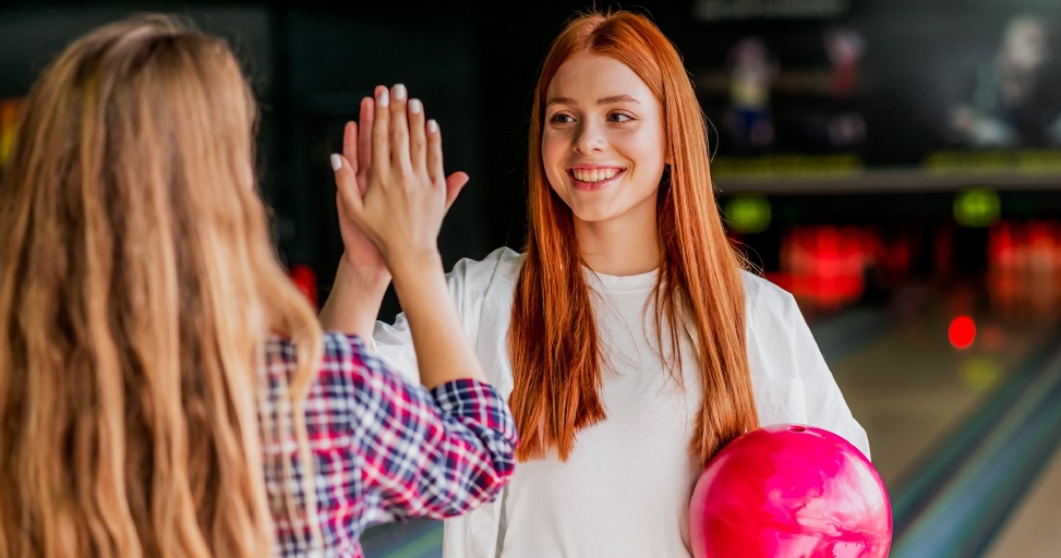 Two young women high-fiving at a bowling alley, one holding a pink bowling ball and smiling.