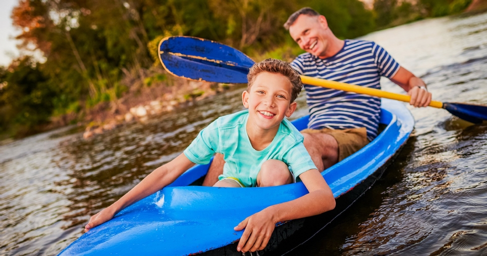 Smiling father and son kayaking together on a peaceful river surrounded by trees.