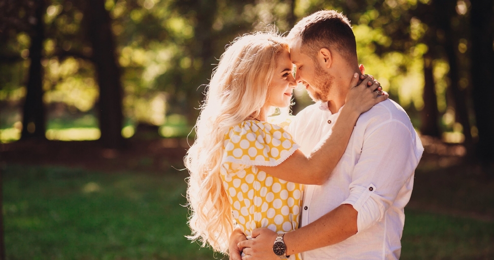 A happy couple embraces in a sunlit park, looking lovingly at each other.
