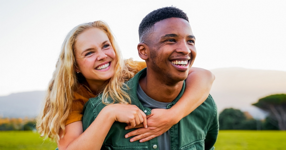 A happy interracial couple smiling broadly outdoors, with the man giving the woman a piggyback ride.