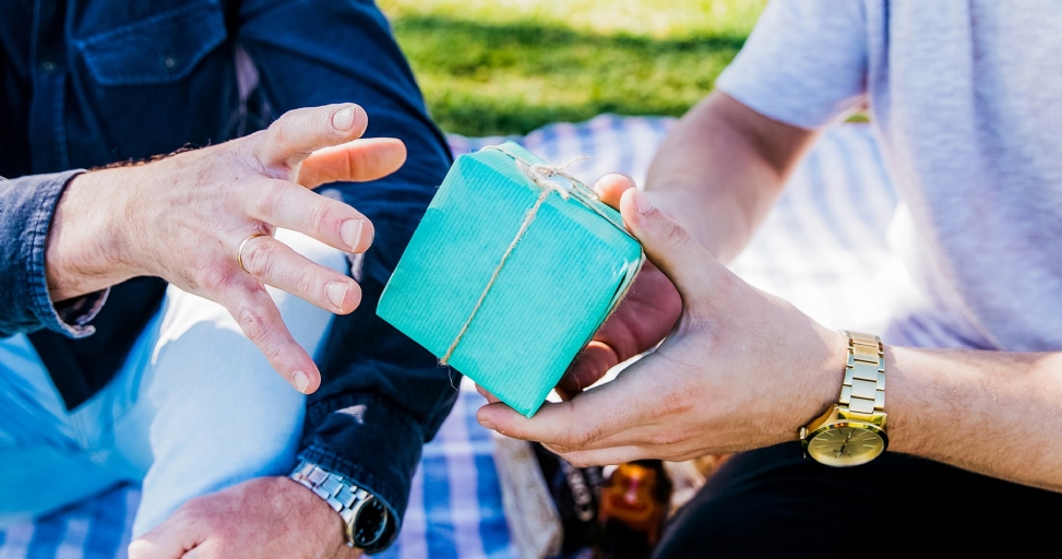 Two people exchanging a teal-wrapped gift outdoors, one wearing a gold watch.