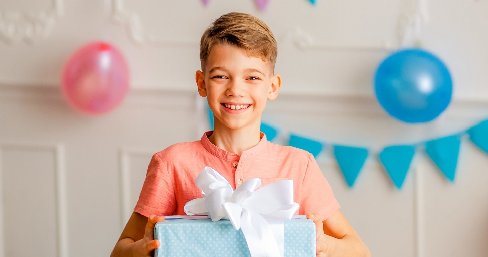 A smiling boy holds a light blue gift box with a white ribbon, surrounded by party decorations.