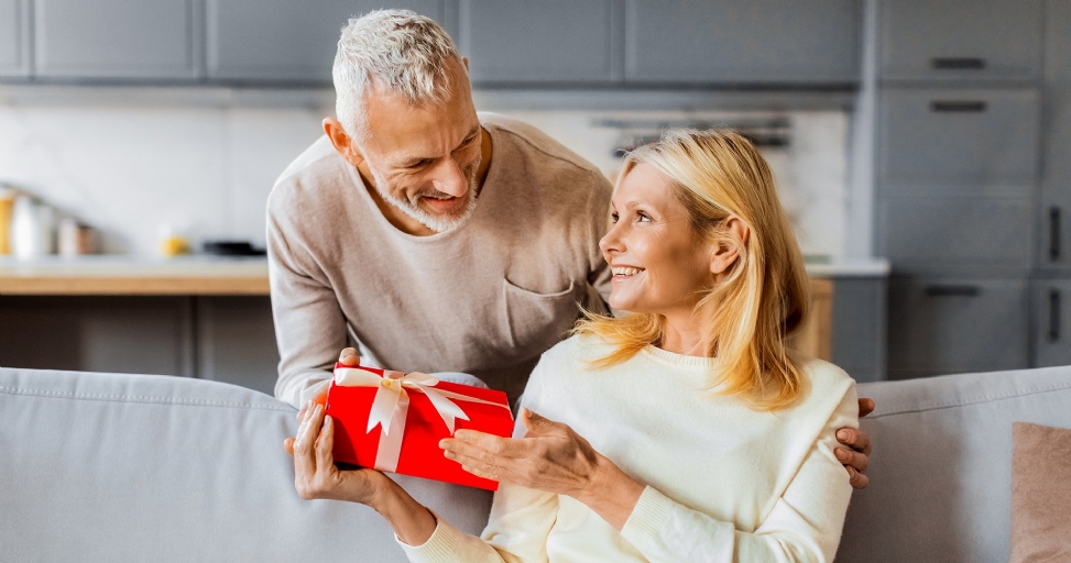 A smiling elderly man gives a red gift box with a white ribbon to an appreciative elderly woman.