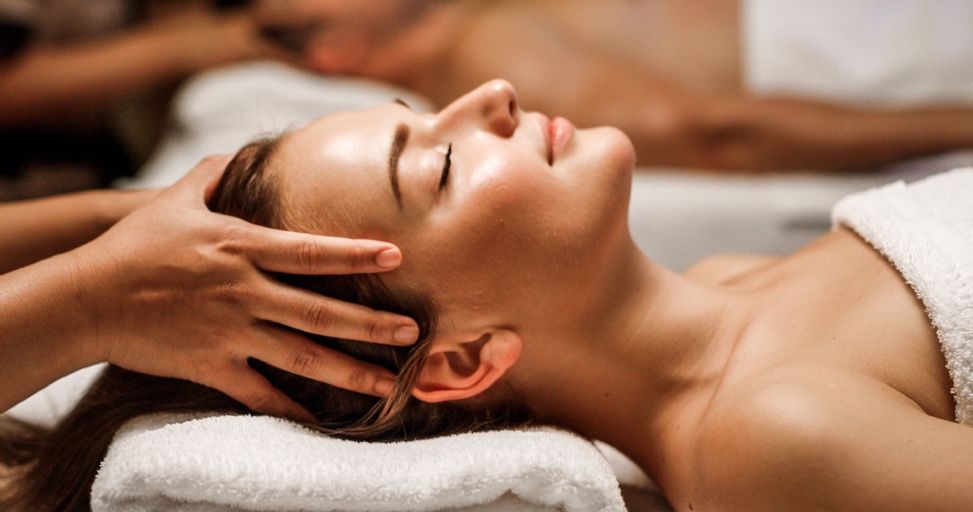 A woman lies relaxed with closed eyes, receiving a soothing head massage in a spa.