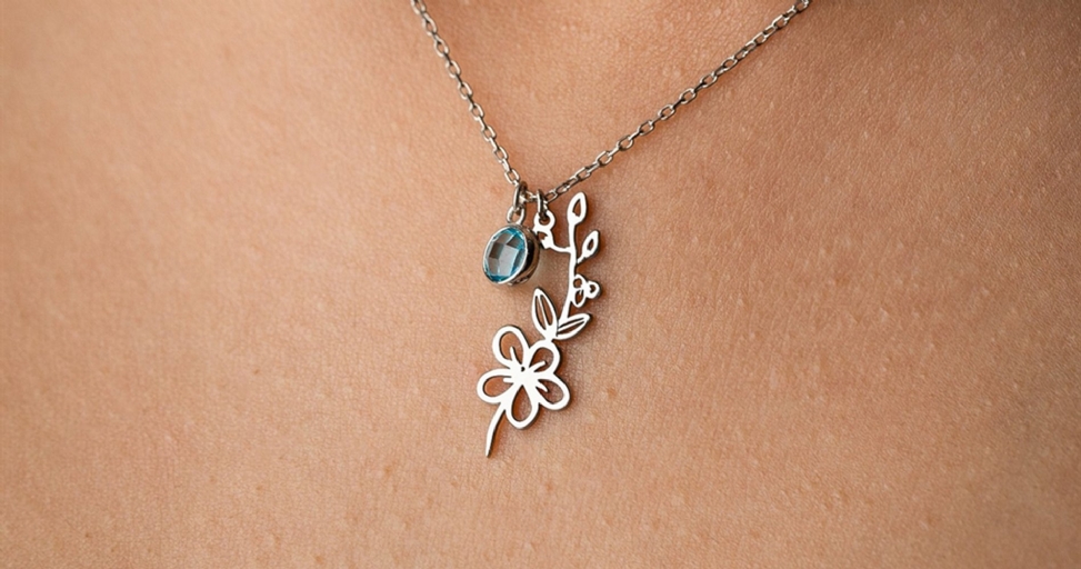 Close-up of a silver necklace featuring an oval blue gemstone and a delicate flower pendant on light skin.