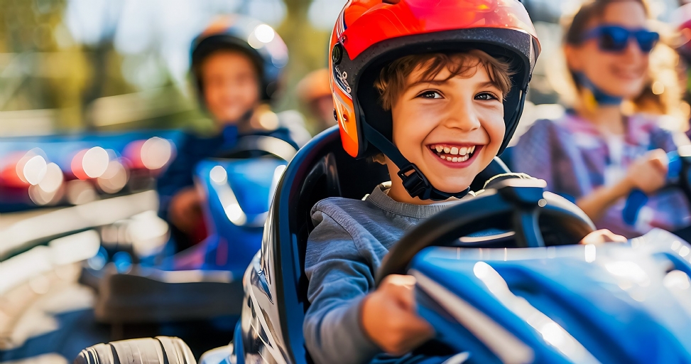 A joyful boy in a red helmet smiles brightly, driving a blue go-kart on a sunny day.